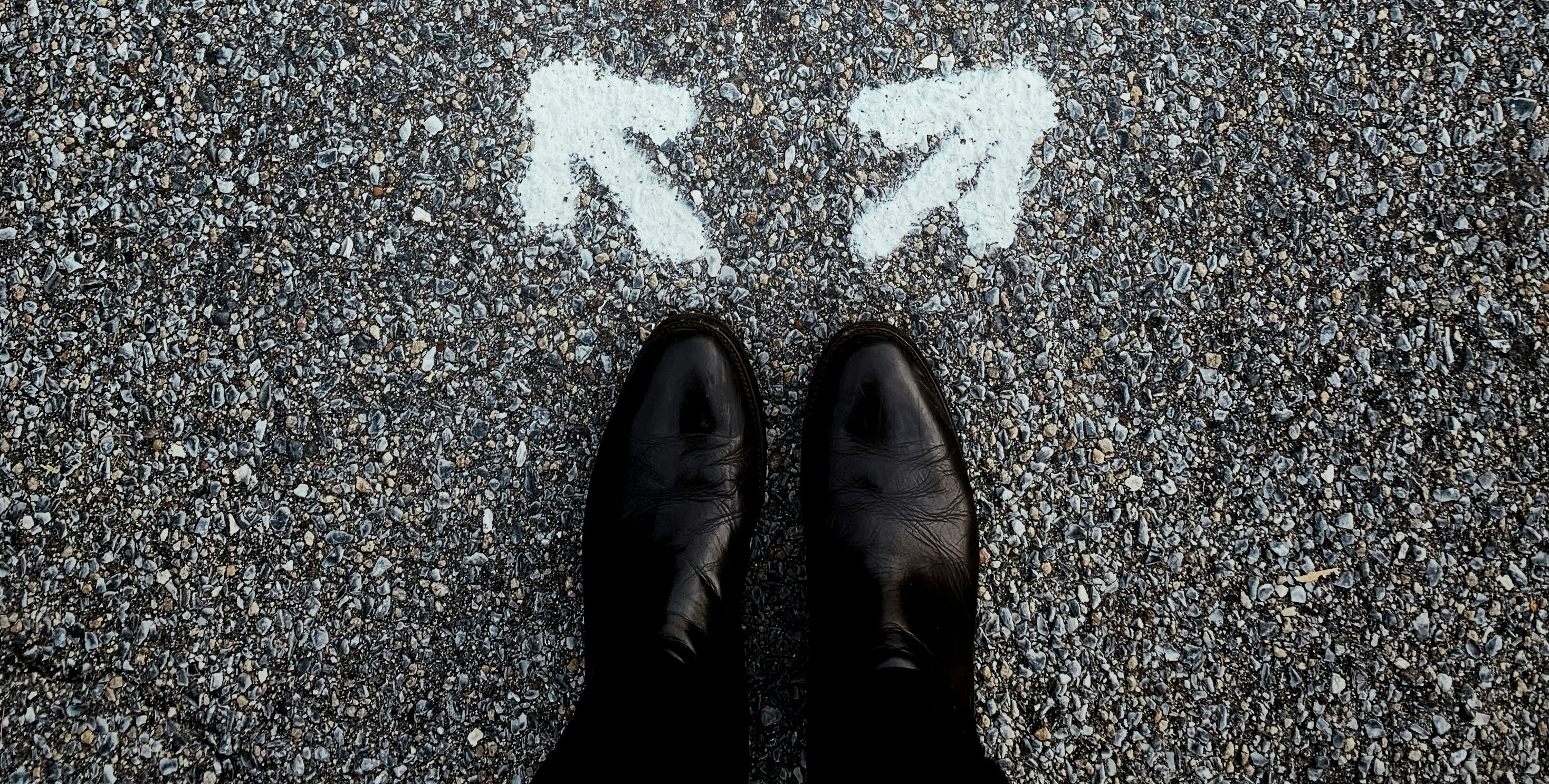 A pair of black shoes standing in front of two white spray-painted arrows pointing in different directions on pavement